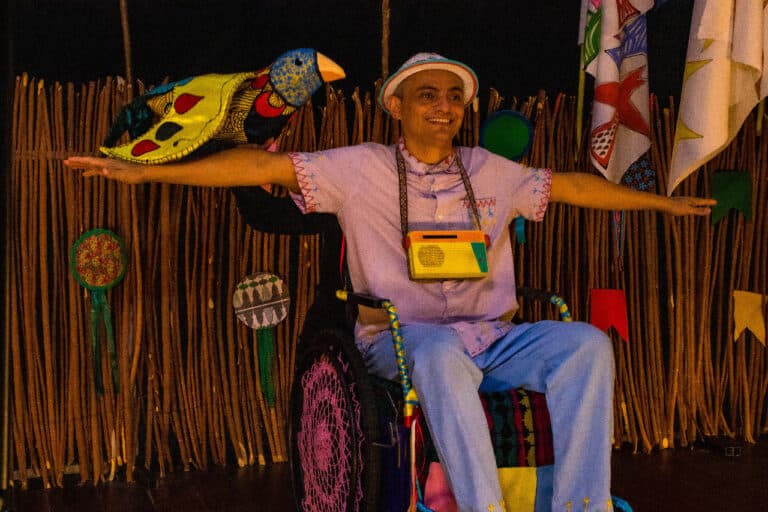 Ator sorridente com os braços abertos e uma marionete de pássaro posa em seu braço, vestindo chapéu e sentado em cadeira de rodas decorada.