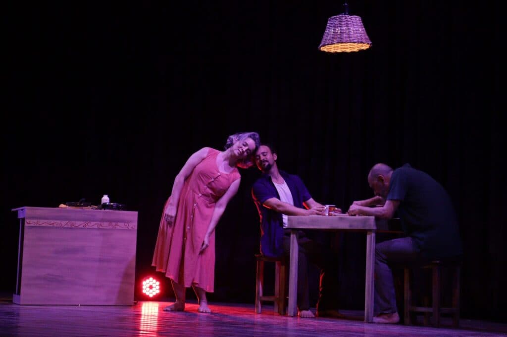 Três pessoas em torno de uma mesa, iluminadas por uma lâmpada pendente em palco escuro.