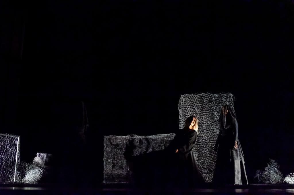 Em um palco escuro, três figuras vestidas de preto estão em cena. Uma mulher de cabelos brancos senta-se, amparada por outra. Ao fundo, painéis de rede cinza compõem o cenário.
